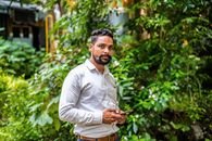 A handsome young man standing outdoors among lush greenery, holding a smartphone.